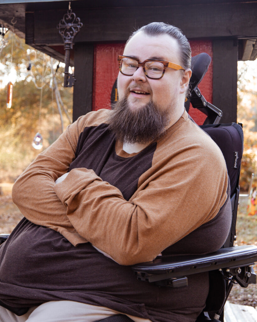 Brentley, sitting in his wheelchair with arms crossed. He is smiling and looking at the camera. Wearing a brown and mustard colored shirt with glasses to match.